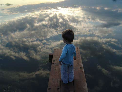 Little boy standing on a small dock staring at the reflection of sky in water.